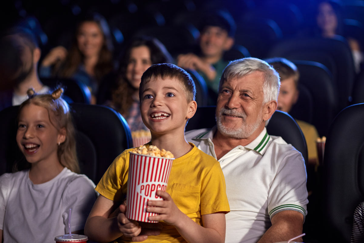 6-year-old white grandson sitting on grandfather’s lap eating popcorn and smiling in a movie theater full of people smiling watching a movie