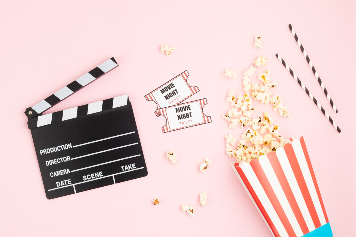 Popcorn bucket, with popcorn spilling out, movie tickets, straws, and clapper board sitting on a pink background. 