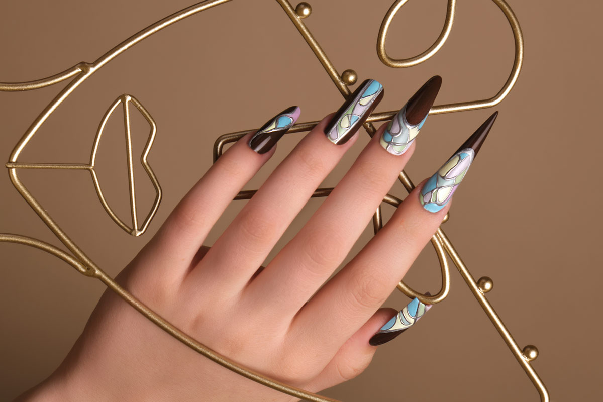 Close-up of a white woman's hands displaying various nail shapes and geometric designs that are brown, light pink, light blue, light yellow, light green, and silver holding a gold wire art piece that is an abstract face.