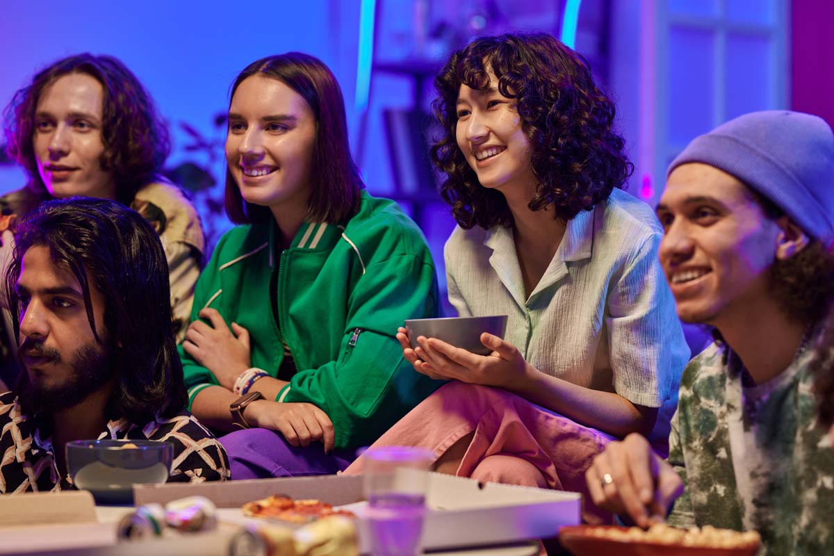 A diverse group of friends in their 30s gathered around a TV screen in a living room, smiling and watching a show, with food on a table in front of them.