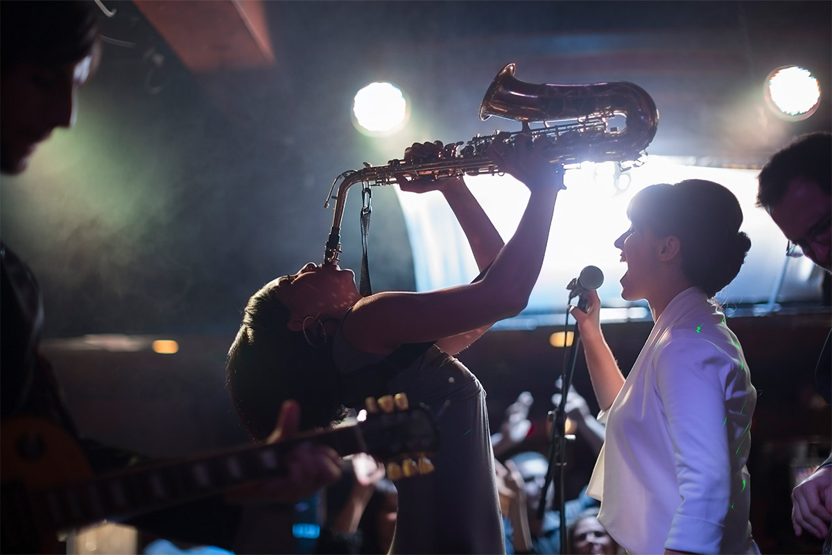 Black woman leaning backwards playing the saxophone passionately with a white woman singing in a club.