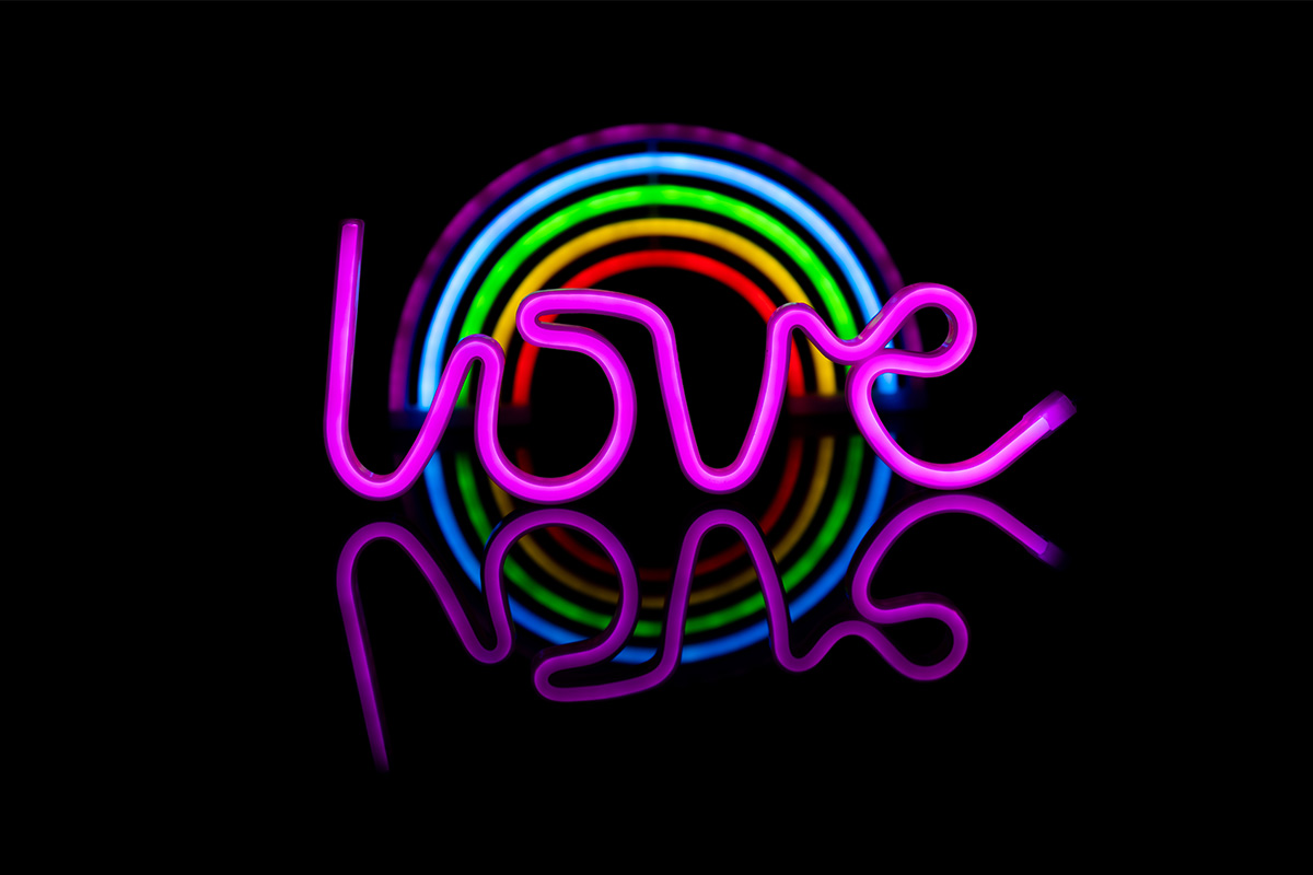 Purple neon cursive “love” sign with neon rainbow sign behind it on a black background.