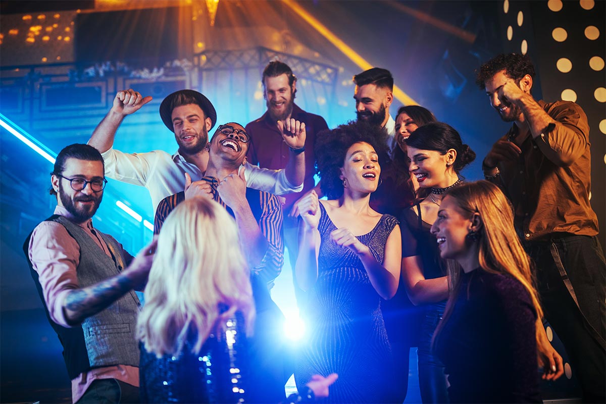 11 multiracial people dancing and laughing in a nightclub with bright lights.