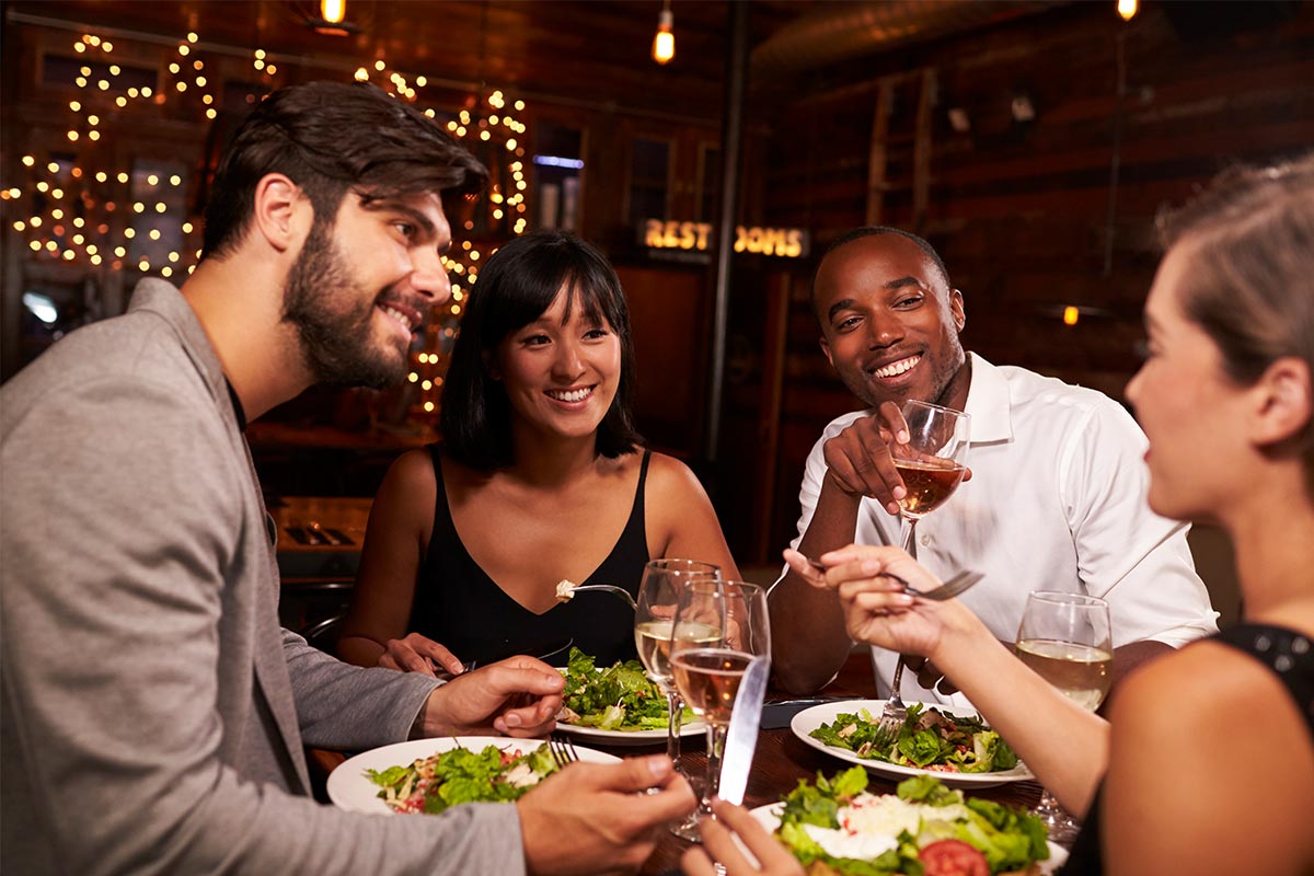 Four multiracial friends eating salad and drinking wine together in a restaurant.