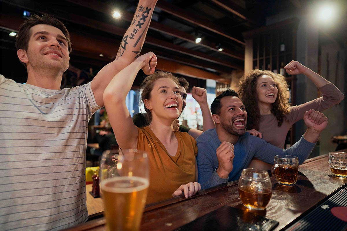 Multiracial group of friends in their 20s to 30s cheering on their team with fists in the air with beer at an indoor sports bar 