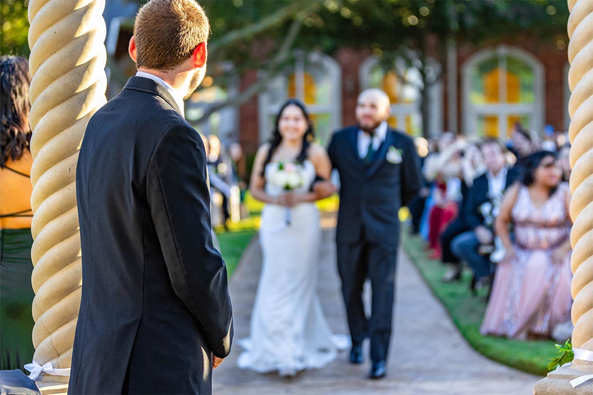 A white groom stands at an outdoor altar as he watches his bride walking down the aisle with her father