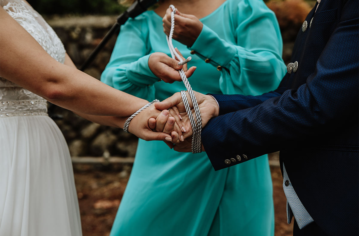 A bride and groom participate in a knot-tying ceremony being performed by a woman in a teal dress.