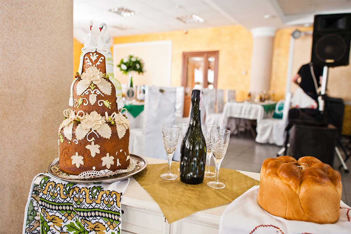 Tall, tiered decorated wedding cake with swan cake topper sits on white and gold table next to champagne and empty champagne glasses with a white and green indoor wedding reception setup in background