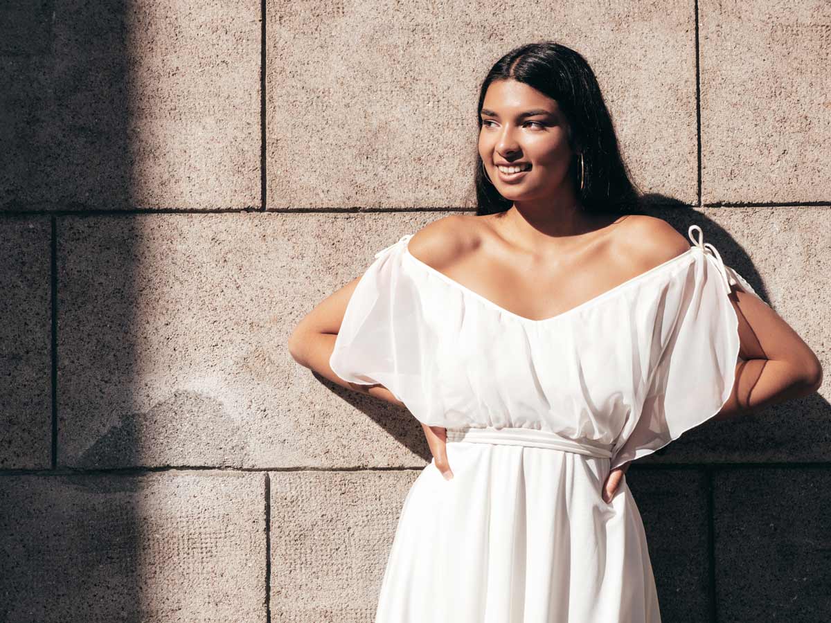 Mixed-race 20-something-year-old woman wearing an off the shoulder white dress leaning against a brick wall. 