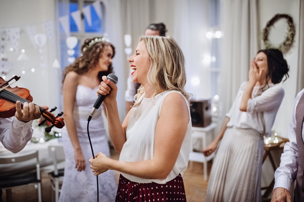 White 35-year-old woman making a funny speech at a wedding reception with guests laughing.