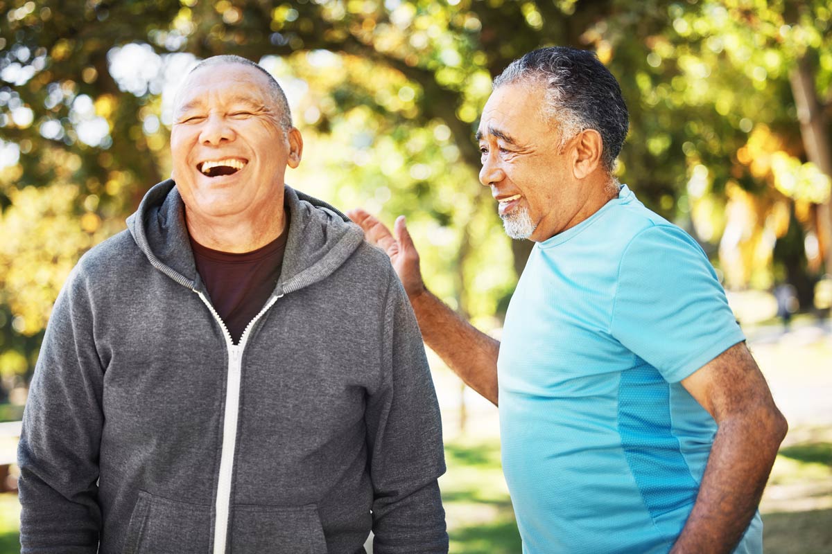 Two older adult brown men stand laughing together outside.