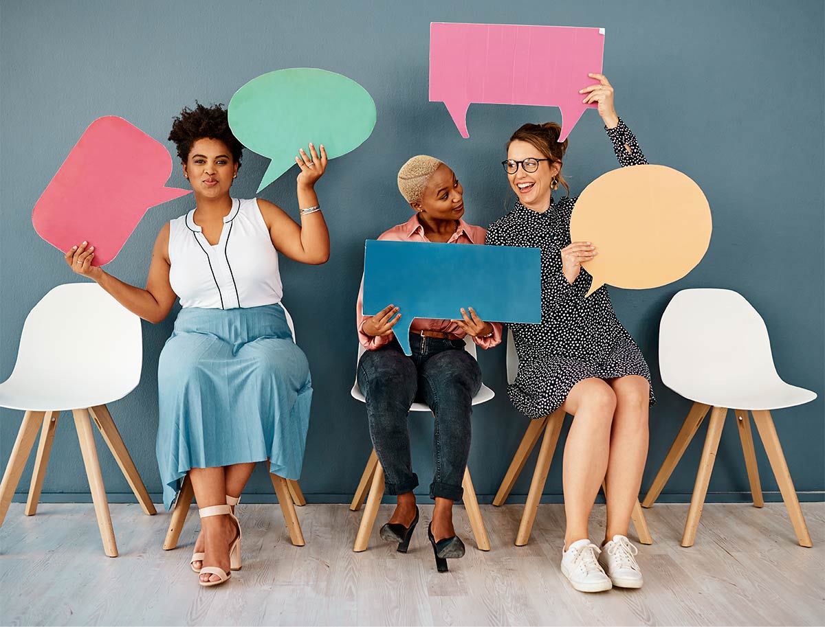 three multiracial women with joyful and amusing expressions sitting on white chairs while holding up colorful paper speech bubble cut outs