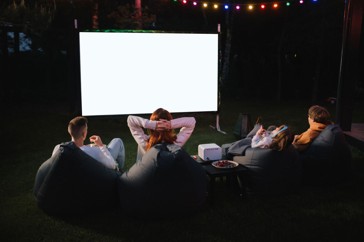 A white family watching a movie projected on a screen outdoors at night sitting on black bean bags.