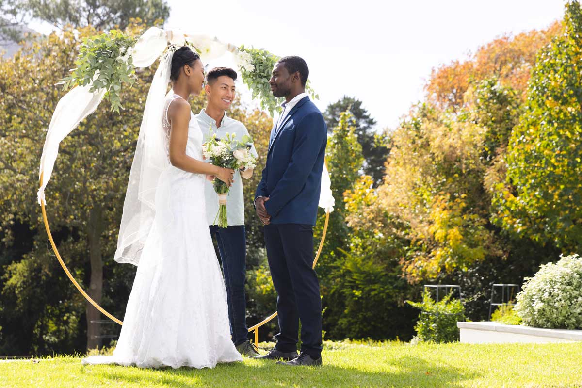 Alt tag: 30 something black bride and black groom standing outdoors under wedding arch with Asian officiant 