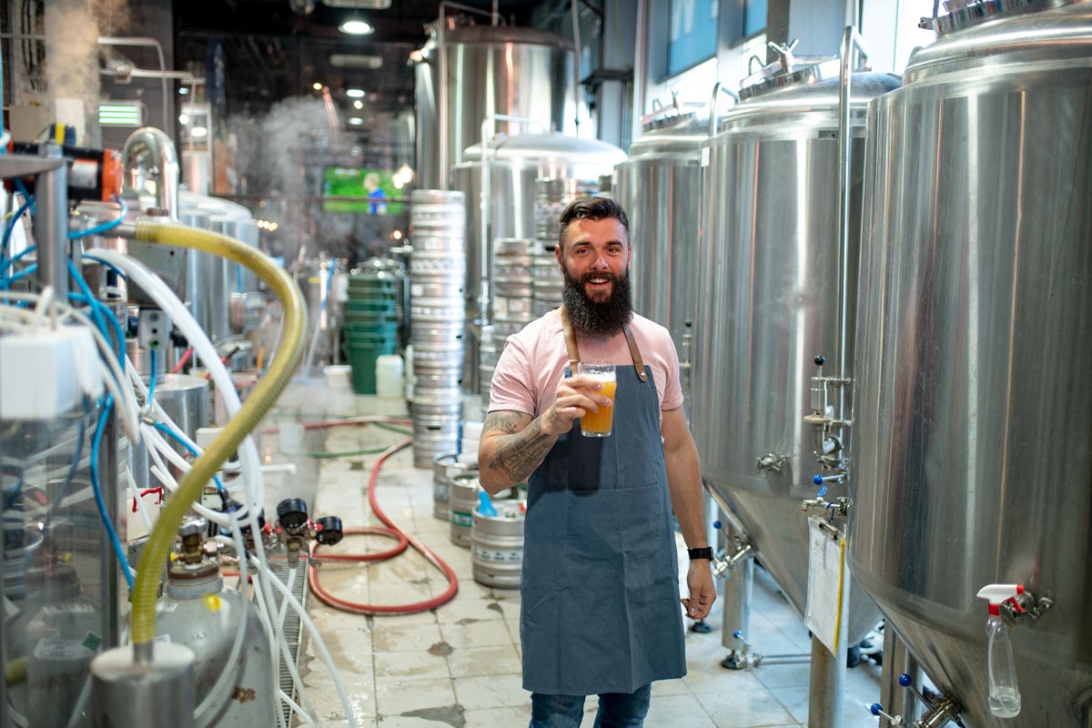 A 35-year-old white man with a beard wearing a pink shirt under an apron taste testing a freshly brewed beer.  