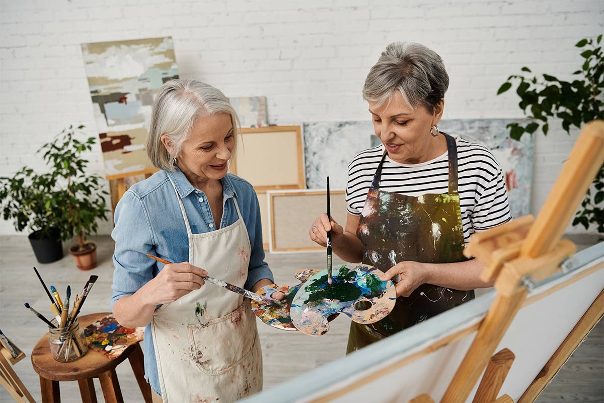 Two white women in their 60s wearing aprons converse as they hold paint brushes and palettes and stand in front of a canvas on an easel in an art studio 