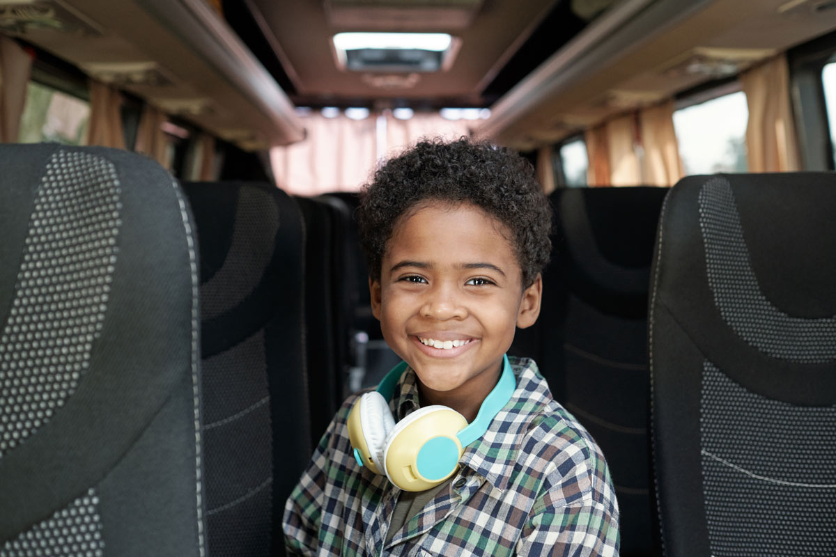  Black 8-year-old boy wearing a plaid button-up shirt and headphones around his neck standing in the aisle of a charter bus. 