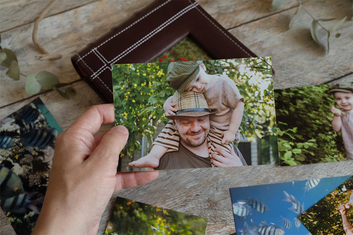 white hand holding a picture of a white father with his son on his shoulders both wearing hats outside with more pictures on the desk in the background