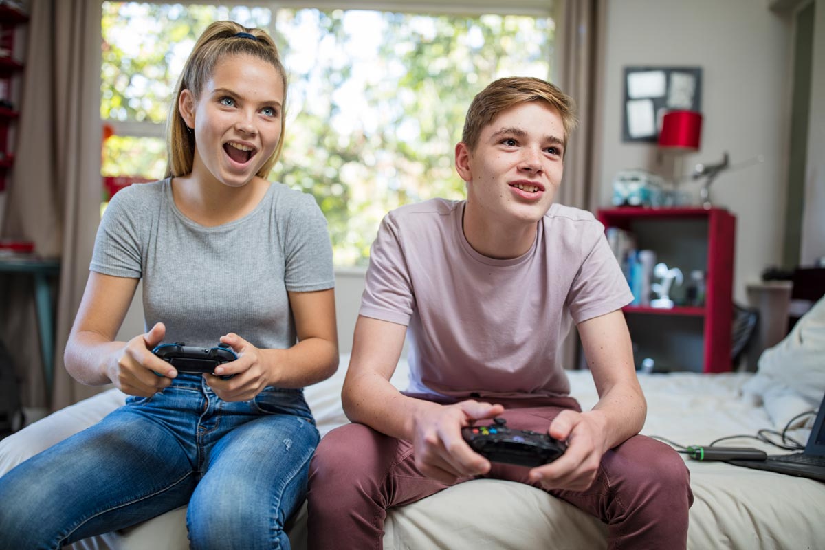 White teenage brother and sister sitting on a bed playing a video game with controllers.