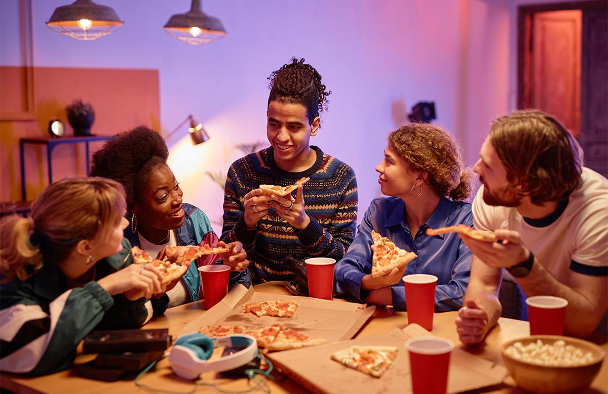 Group of five multiracial adults in their 30s share pizza and drink from red cups while talking and smiling around a table indoors