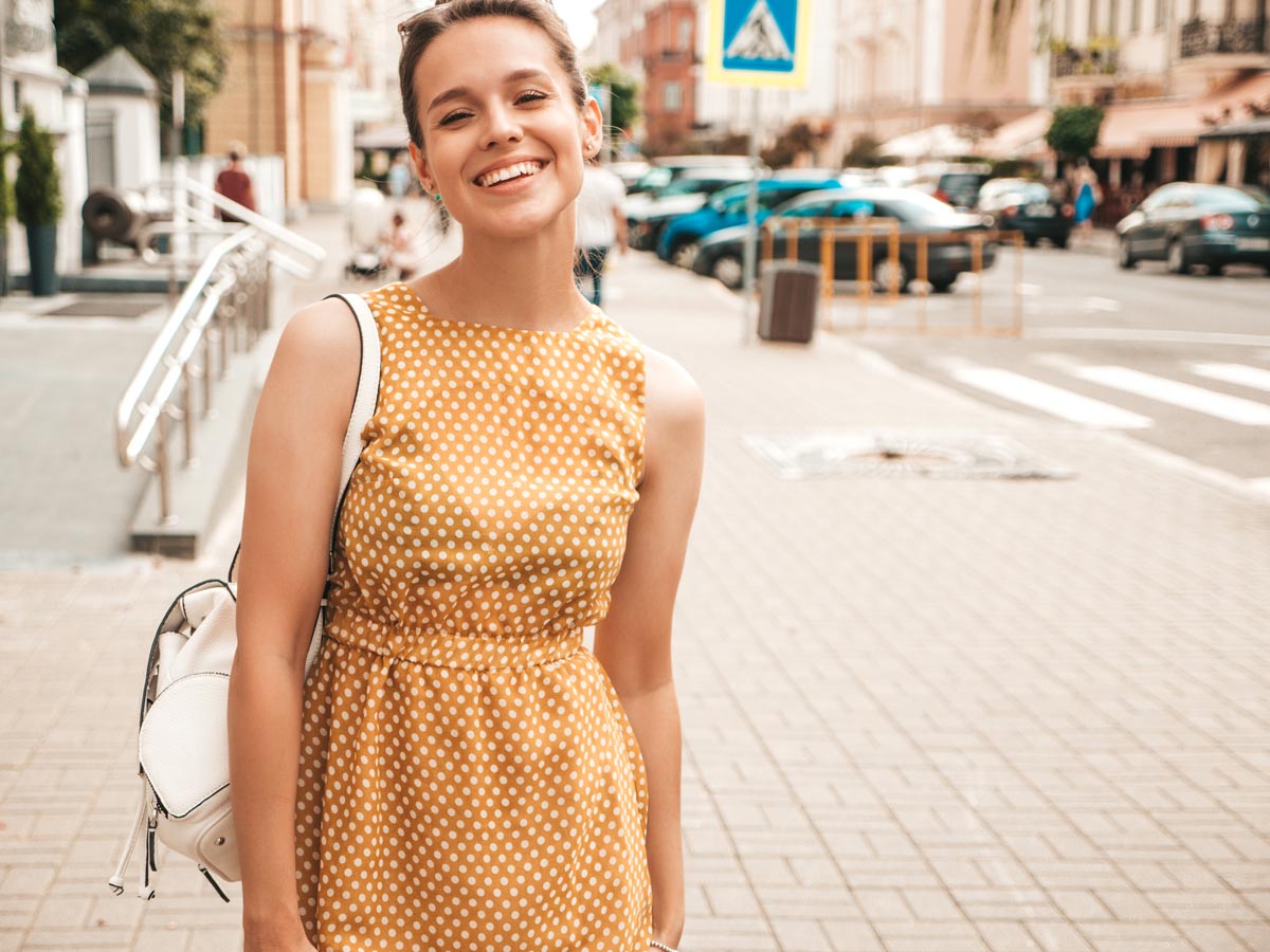 White 25-year-old woman smiling and wearing an orange polka dot dress. 