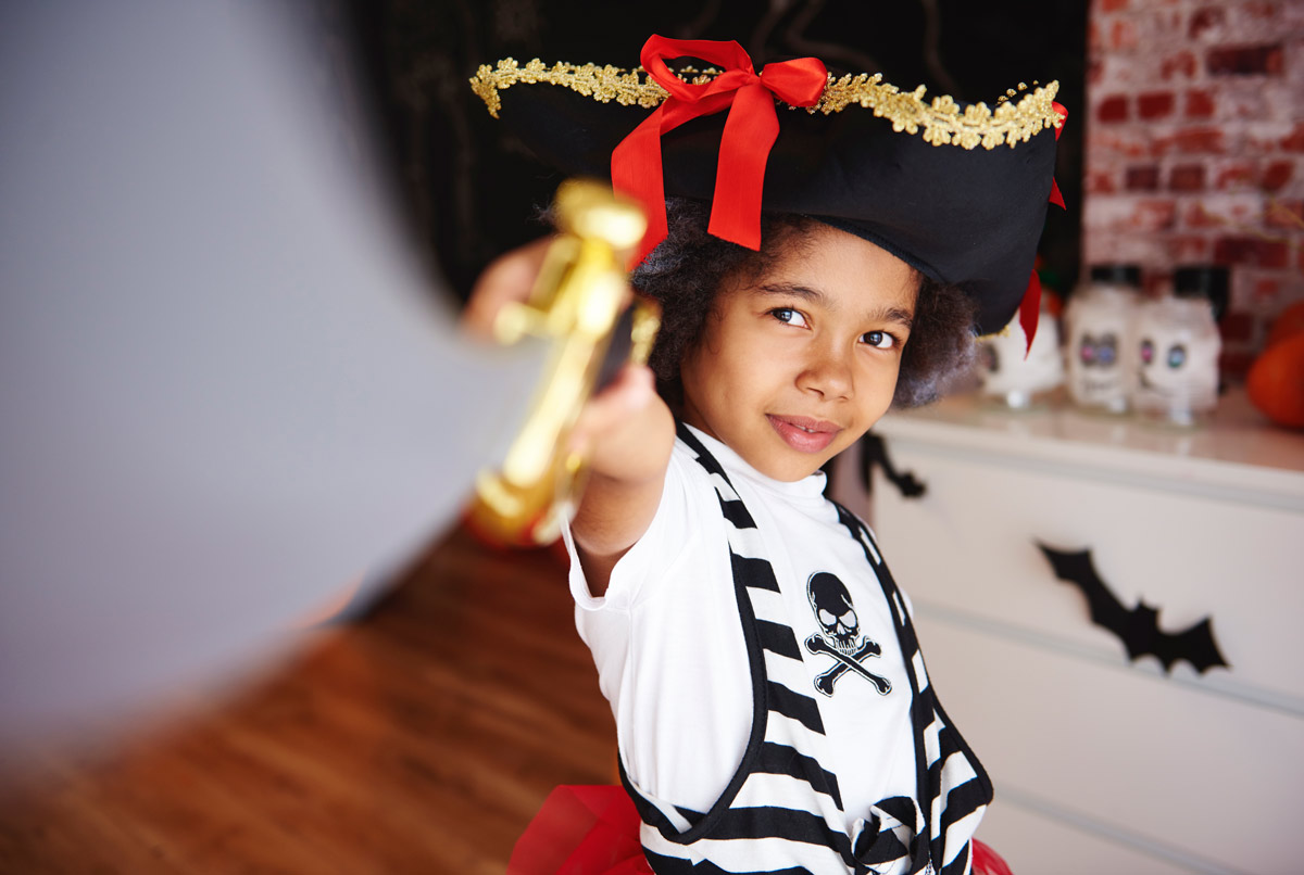 Black child wearing a pirate costume and waving a plastic sword at the camera in the living room