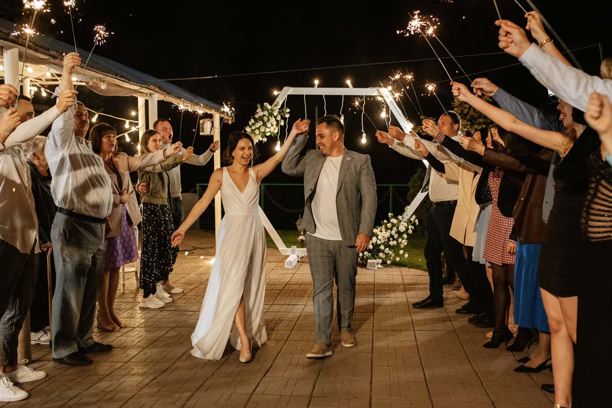 White 30 something woman in wedding dress and hispanic 30 something man in gray suit hold hands and walk down path from altar outdoors at night surrounded by people holding sparklers 