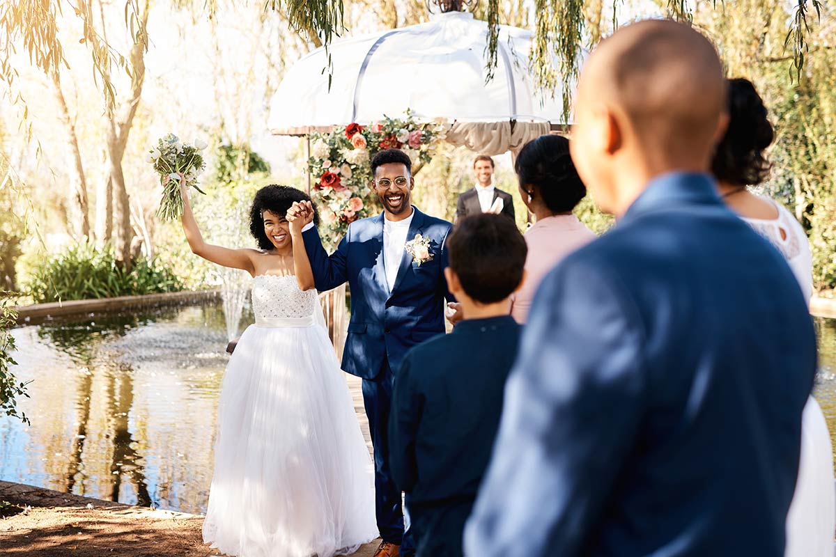 Black 30 something married couple cheering with smiles and arms in the air as they walk hand in hand down outdoor wedding aisle with flowers and water in the background