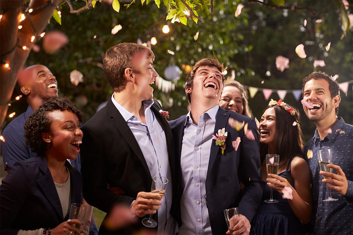 Two white men in their 30s smile and celebrate marriage with multiracial friends in their 30s and champagne outside at night while pink flower petals fall around them