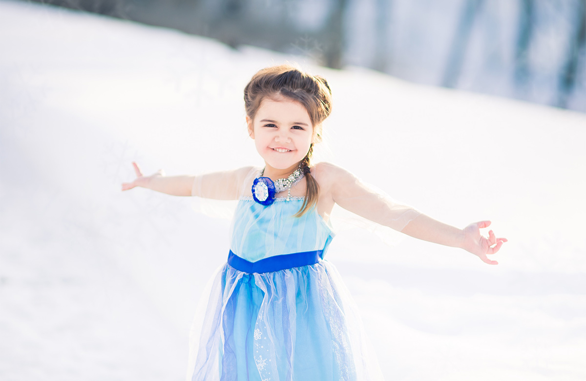 white little girl, in an ice princess costume standing outside during winter, with her hands out by her side and smiling. 