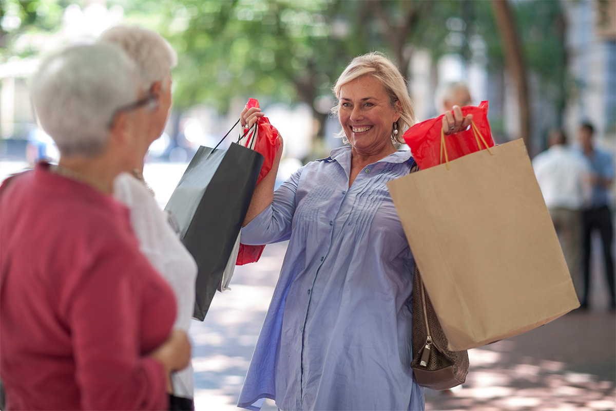 White woman in her 50s to 60s wearing light blue blouse outside during sunny day smiles and holds up multicolored shopping bags as she faces two other blurred women