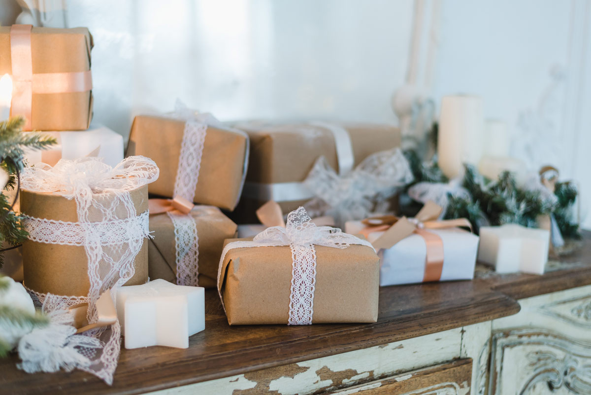 Neatly stacked white, brown, and bronze wrapped gifts with bronze and white lace ribbons on a brown and white wooden table with candles and plants for decoration