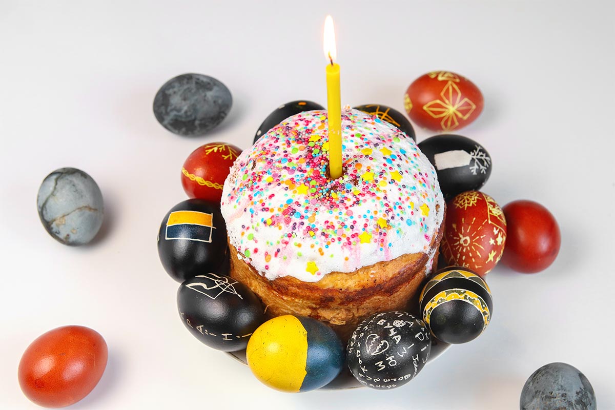 Vanilla cake with white icing on it with colorful sprinkles and a yellow burning candle with red, grey, black, and yellow easter eggs surrounding it