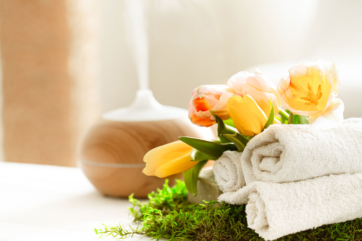 Spa closeup with aromatherapy essential diffuser and towels with yellow and peach flowers on top of fake grass