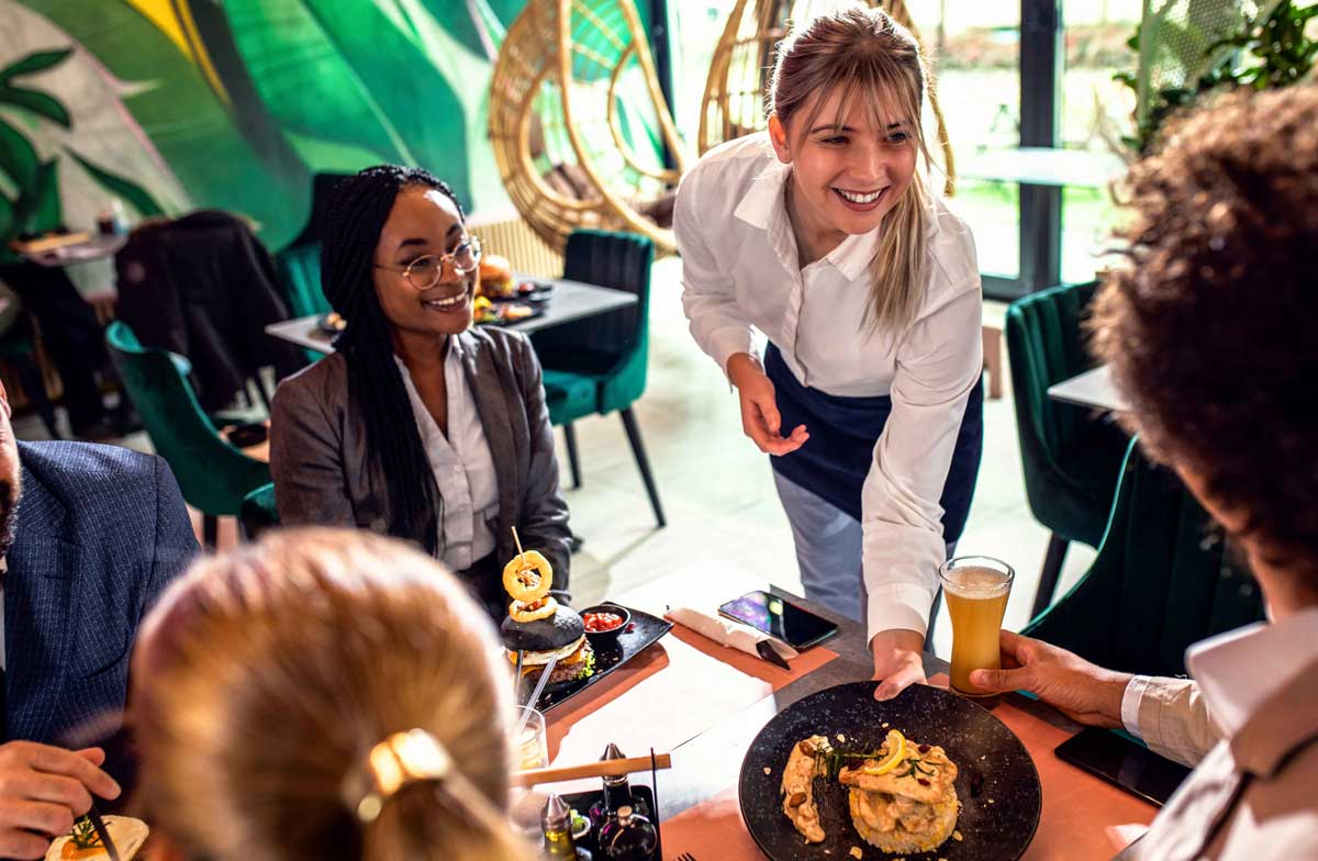 Young adult white waitress serving entree with a smile to table of 4 adult multiracial guests at an indoor green restaurant 