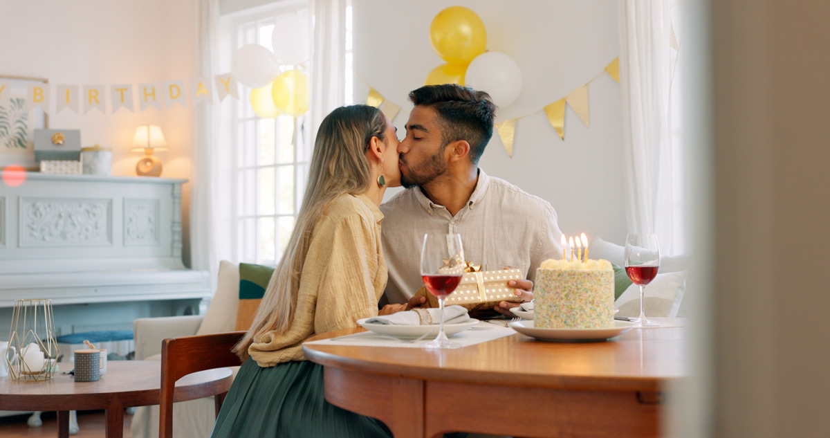 Hispanic man and white woman in their 30s kiss on the cheek while sitting at a table with wine birthday cake and decorations