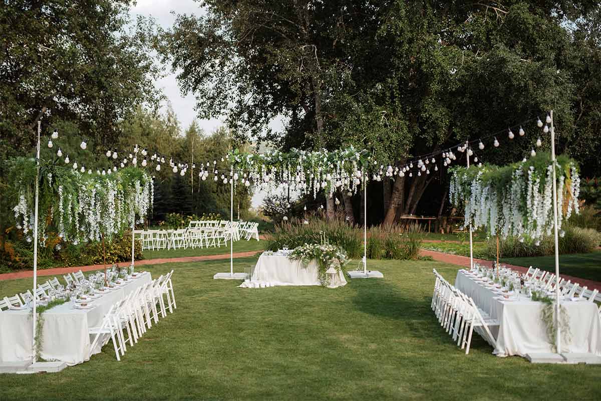  A rustic garden wedding with string lights, tables with white table cloths, and decorative flowers hanging from poles.