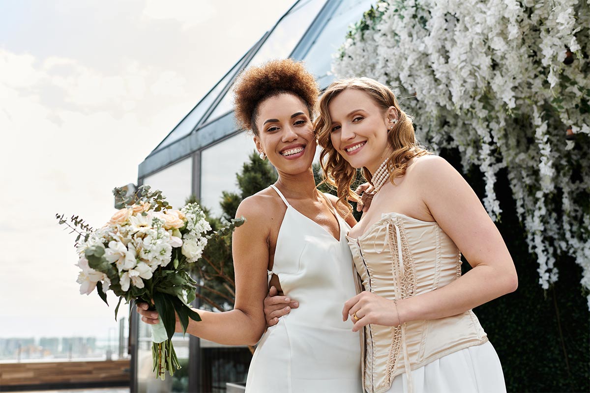 White 20-something woman in wedding dress hugs black 20-something woman in wedding dress holding bouquet as they smile at the camera in front of a botanical garden covered in white hanging florals