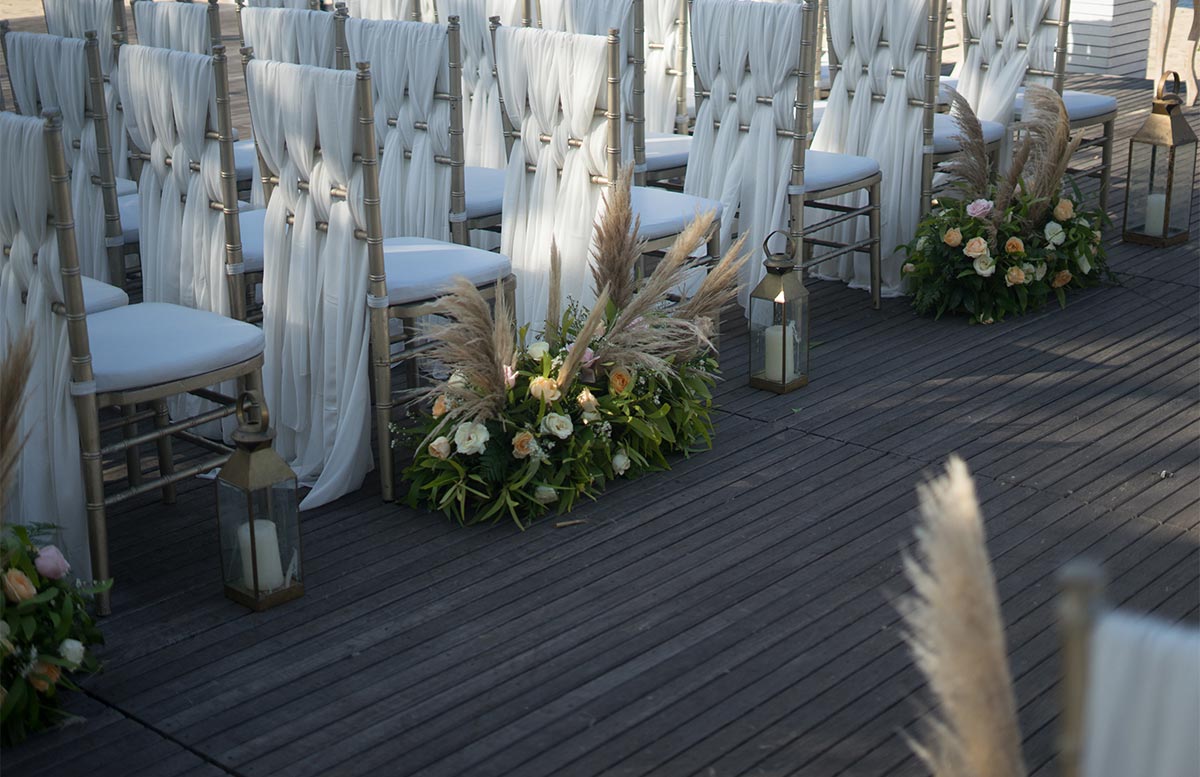 white wooden chairs lined up on a wooden deck, with white cloth hanging off the back of them and the chairs are next to gold lanterns and flower arrangements