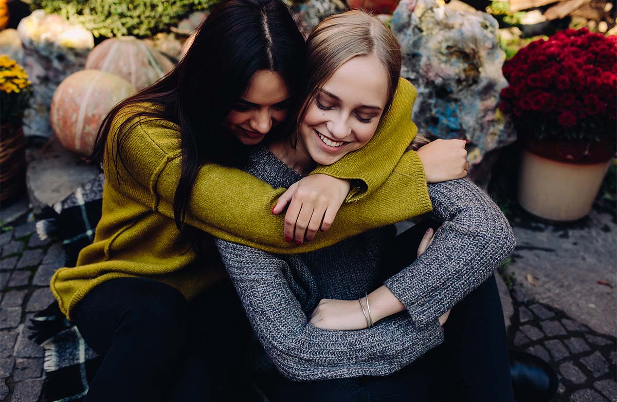 20-something white adult female hugging her 20-something white friend from behind as they both smile and sit on stone path outside