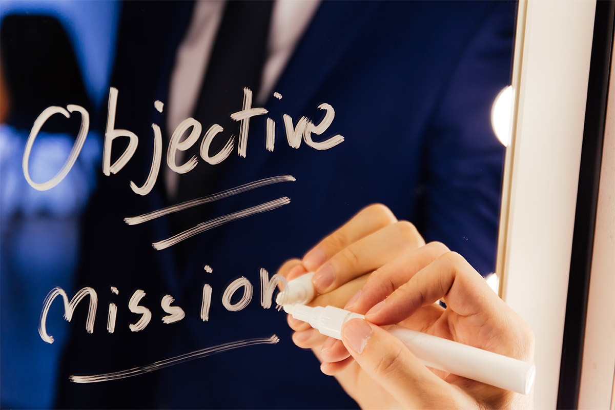 White hand, writing “objective mission” on a mirror with a white marker with a suit and tie in the reflection.