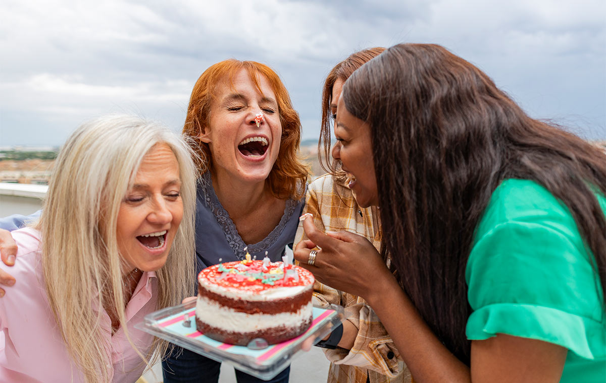 Group of multiracial women in their 50s-60s hug and laugh together around a birthday cake while outside during the day  