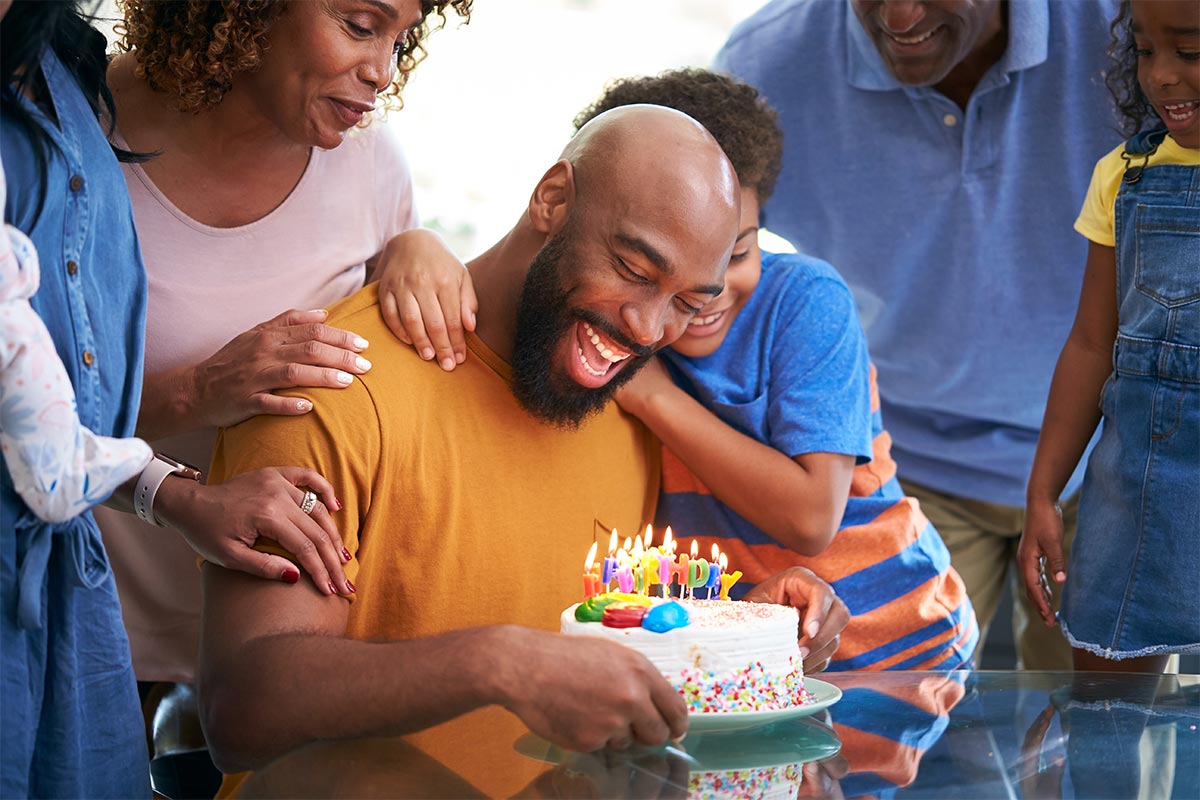 Black man in his forties sitting at table to blow out his birthday candles laughs while embraced by his son and other surrounding family members