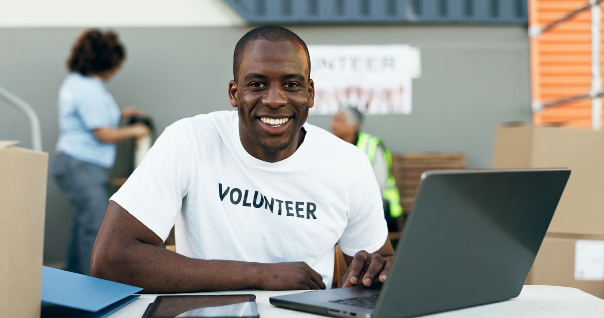 A black 25 year old man sits at a desk smiling wearing a shirt that says volunteer.