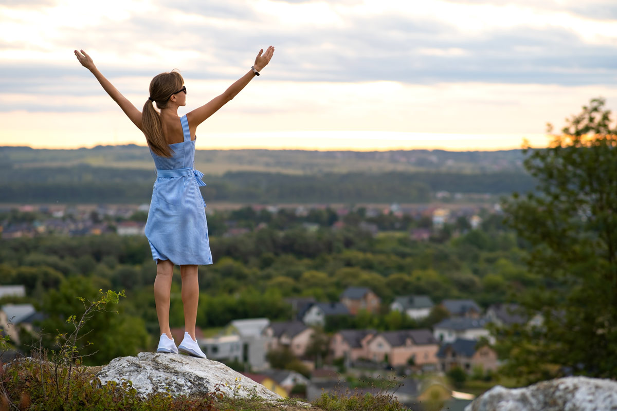 Woman in blue striped dress holding hands in the air at the top of a rocky cliff