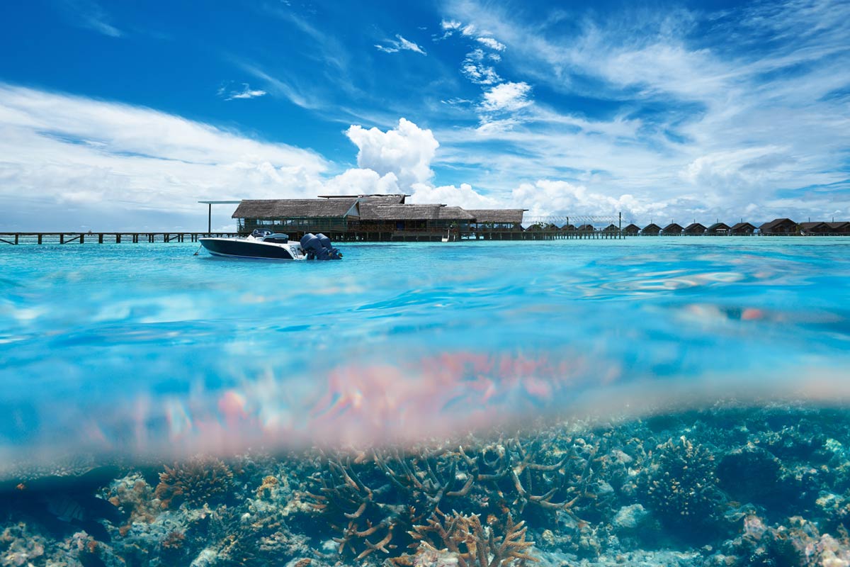 Small boat sits in clear blue water above coral reefs on a sunny day while a pier and building sit in the background