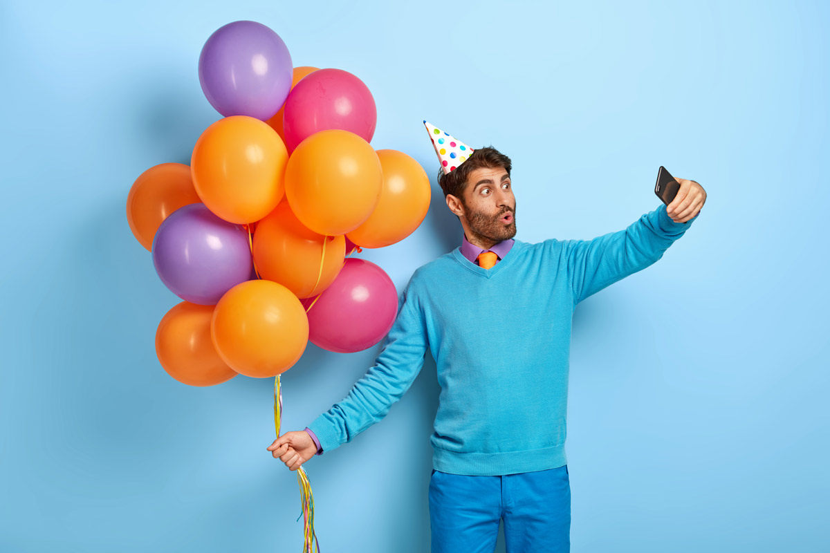 A white man in a birthday hat holding balloons takes a selfie.