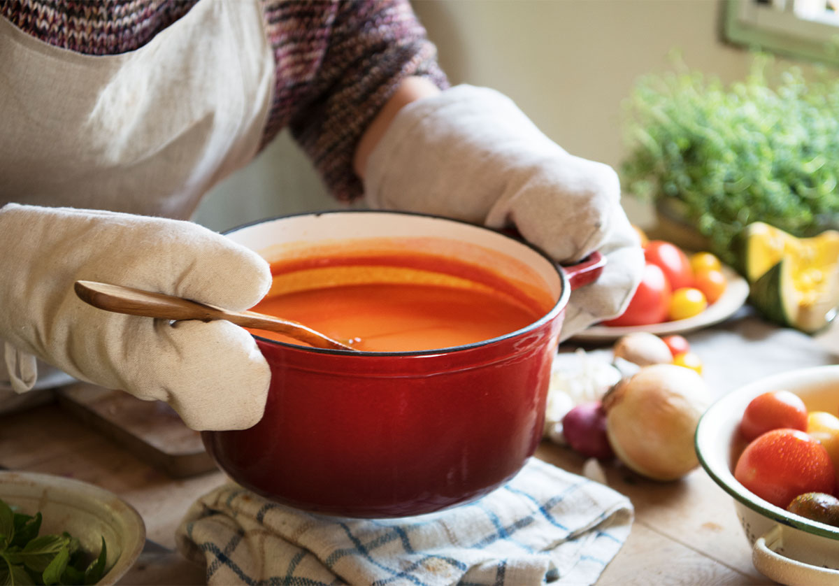 woman wearing gloves and an apron holds a big red bowl of tomato soup above a table that has a towel and numerous vegetables on it