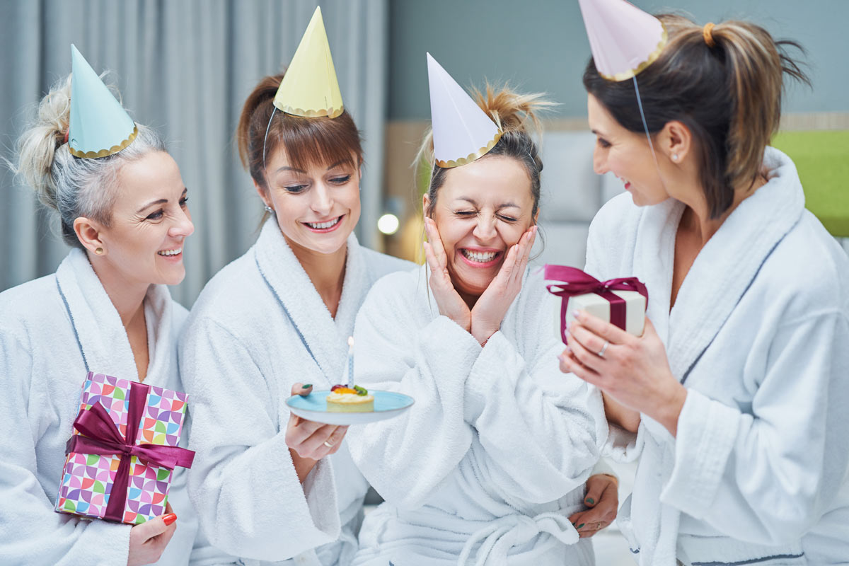 4 White 20 something women wearing white robes and birthday hats, holding presents, and a cupcake with a candle. 