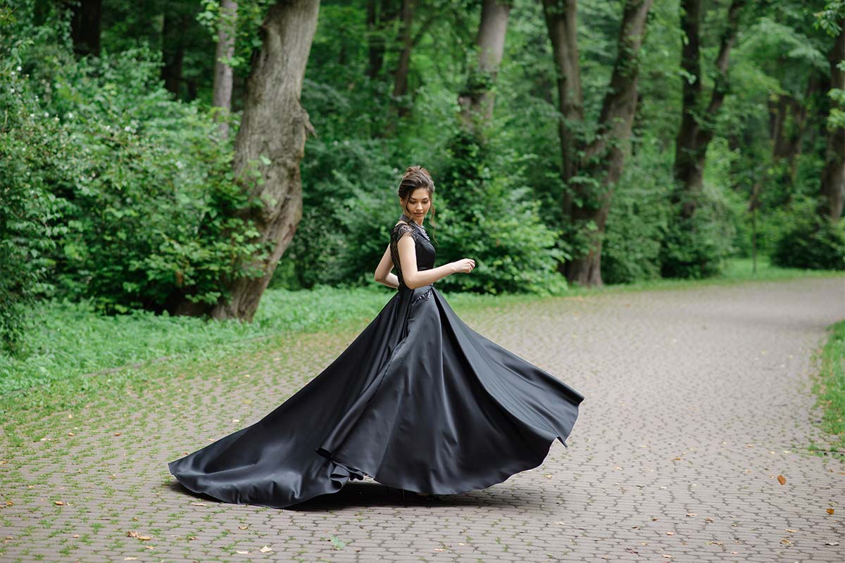 White bride in her 20s twirls outside on stone path by dense trees in black wedding dress